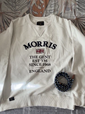 Vit sweatshirt från Morris - Vit sweatshirt från Morris med broderad text och brittisk flagga på bröstet. Rund halsringning, ribbade muddar och klassisk passform. Perfekt för en stilren och avslappnad look. Skick 8.5/10 priset ej hugget i sten🤩 hör av er vid minsta lilla fundering 😇