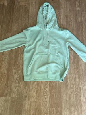 Mintgrön hoodie med magficka - En stilren mintgrön hoodie med huva och dragsko. Hoodien har en stor magficka framtill och ribbade muddar vid ärmslut och nederkant. Perfekt för en avslappnad look. Materialet känns mjukt och bekvämt mot huden.