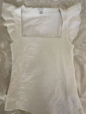 Vit blus med volangärmar från Amisu - Supersöt vit blus från Amisu med fyrkantig ringning och volangärmar. Tunt, lätt material med diskret struktur som ger en elegant känsla. Perfekt att styla med jeans eller kjol för en fräsch look.