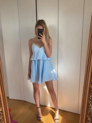  Ny Ljusblå kort klänning med volang - Söt ljusblå kort klänning med volangdetalj och smala axelband. Klänningen har v-ringning och en luftig passform som ger en avslappnad och trendig look. Perfekt för varma dagar och somriga tillfällen. Stå L på etiketten men passar medium 