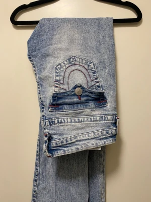 Ljusblå slitna jeans från True Religion - Säljer ett par ljusblå jeans från True Religion med slitningar och hål på benen. Jeansen har klassiska röda sömmar, unik bakficka med lock och knapp samt en snygg tvättad look. Perfekta för en avslappnad streetstil. En knapp har tyvärr lossnat.