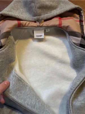 Burberry hoodie  - Snygg grå hoodie från Burberry med dragkedja och klassiskt rutigt foder i huvan. Mjuk insida och praktisk huva med snörning. Perfekt för dig som gillar stilrena och exklusiva plagg. Materialet känns mjukt och bekvämt.