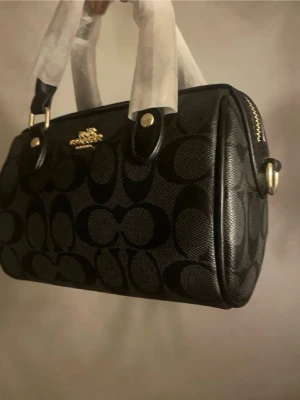 Coach - Snygg svart handväska från Coach med klassiskt monogrammönster i svart och mörkgrått. Väskan har guldfärgade metalldetaljer, dubbla handtag och dragkedja upptill. Tillverkad i tåligt konstläder med struktur. Perfekt för dig som gillar stilrena accessoarer.