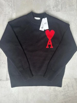 Svart stickad tröja från Ami Paris - Svart stickad tröja från Ami Paris med röd hjärtlogga och bokstaven A på bröstet. Tröjan har rund halsringning och ribbade muddar vid ärmslut och nederkant. Tillverkad i ekologisk bomull enligt GOTS-standard.