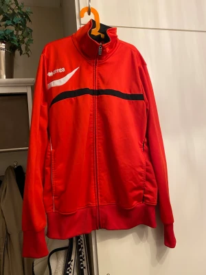 Retro träningsjacka, Erreá - Retro träningsjacka från Errea med dragkedja, ståkrage och svarta samt vita detaljer över bröstet. Jackan har ribbade muddar och två fickor framtill. Perfekt för träning eller sportiga outfits.