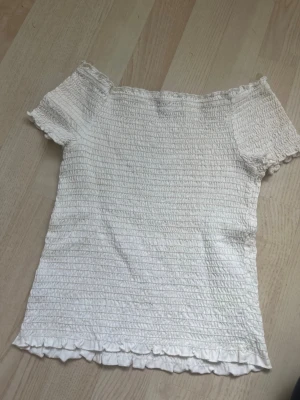 Vit smockad topp med volangkant - Vit stretchig topp med korta ärmar som går att ha off shoulder om man vill. Den passar mig som är xs! Superfin sommartopp som går att styla till allt.