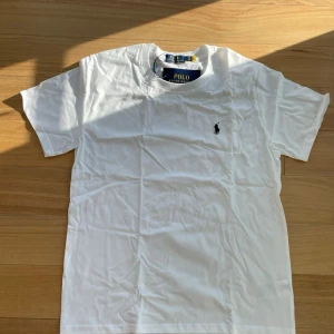 Vit Ralph Lauren T-shirt - 👕 Märke: Polo Ralph Lauren 📏 Storlek: L 🎨 Färg: Vit med svart logga ✨ ☀️ Perfekt för sommaren – stilren och fräsch t-shirt i mjuk bomull från Polo Ralph Lauren. Klassisk passform med ikonisk svart logga på bröstet. Passar perfekt till både shorts och jeans.  ✨ Skick: Helt ny med tags kvar 🧼 Aldrig använd 👌 Tidlös och snygg basic som funkar till allt