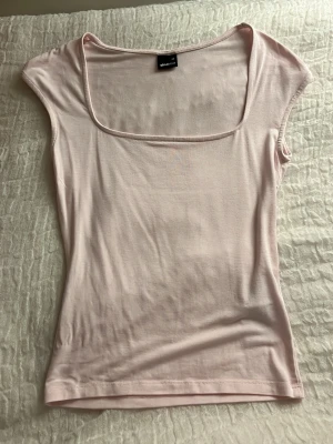Ljusrosa topp från Gina Tricot - Snygg ljusrosa topp från Gina Tricot med fyrkantig ringning och korta ärmar. Tillverkad i mjukt och stretchigt material som sitter skönt mot kroppen. Perfekt att styla med jeans eller kjol för en trendig look.