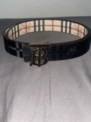 B*B svart bälte dubbel - Snyggt svart bälte från B*B med klassiskt rutigt mönster och ikoniskt TB-spänne i metall. Insidan har B*B signaturrutor i beige och svart. Perfekt accessoar för att lyfta din outfit.