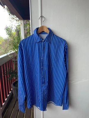 Blåvit randig skjorta med knappar - Snygg blå skjorta med vita vertikala ränder och klassisk krage. Skjortan har långa ärmar och knäppning framtill. Perfekt för en stilren look och passar till många olika outfits.