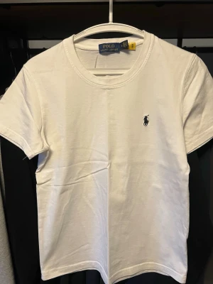 Vit t-shirt från Polo Ralph Lauren - Klassisk vit t-shirt från Polo Ralph Lauren med korta ärmar och rund halsringning. T-shirten har en liten broderad logga på bröstet och är tillverkad i mjuk bomull för en skön känsla. Perfekt basplagg till garderoben.