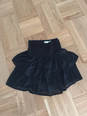 Svart volangkjol från Kappahl - Supersöt svart kjol från Kappahl med bred smock i midjan och dubbla volanger. Kjolen har en luftig och flowy känsla, perfekt för att skapa en cool och trendig look. Passar dig som gillar att sticka ut lite extra!