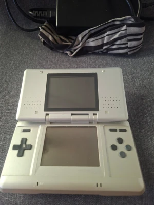 Nintendo DS silver bärbar spelkonsol - Nintendo DS i silver med dubbla skärmar och klassiska knappar. Konsolen är kompakt, vikbar och tillverkad i plast. Perfekt för portabel gaming och retrospel. En ikonisk handhållen konsol från Nintendo med stilren design.