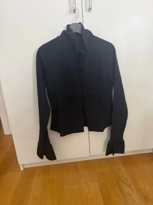Svart skjorta med vida ärmslut - Stilren svart skjorta med lång ärm. Skjortan har knappar framtill och en krage som ger en cool och modern look. Perfekt för dig som gillar att sticka ut med detaljer.