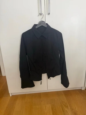 Svart skjorta - Stilren svart skjorta med klassisk krage och knäppning framtill. Skjortan har långa ärmar. Skjortan har korsett knäppning baktill som ger en cool och modern look. Materialet känns mjukt och bekvämt mot huden. Köpt second hand i nyc.