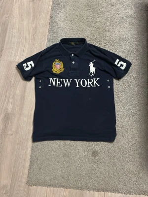 Mörkblå pikétröja Polo Ralph Lauren - Snygg mörkblå pikétröja från Polo Ralph Lauren med vit broderad logga, texten NEW YORK framtill och siffran 5 på ärmarna. Tröjan har krage, knappar och detaljerade broderier i gult och vitt. Perfekt för en sportig och stilren look.