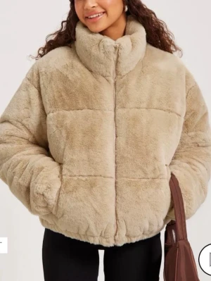 Beige teddyjacka med hög krage - Mysig beige teddyjacka med hög krage och dragkedja framtill. Jackan har en boxig passform och är tillverkad i mjukt, fluffigt material som ger en trendig look. Perfekt för kalla dagar och enkel att styla med olika outfits.