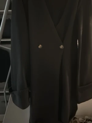 Svart Adoore klänning med knappar - Stilren svart Adoore klänning med djup v-ringning och dubbla knappar framtill. Klänningen har långa ärmar med uppvikta muddar och en rak passform. Perfekt för en trendig och chic look.