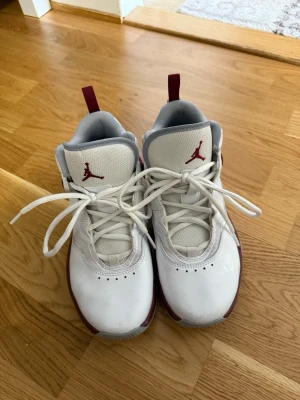Vita och vinröda Air Jordan sneakers - Snygga Air Jordan sneakers i vitt och vinrött med klassisk Jumpman-logga på plösen. Skorna har högt skaft, vita snören och grå insida. Ovandelen är i en mix av läder och textil, med detaljer i vinrött längs sidorna och sulan. Perfekta för dig som gillar streetstyle och basket.