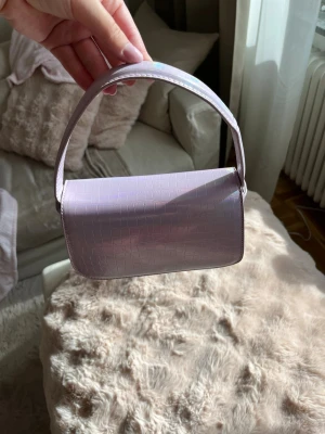 Ljuslila croco handväska - Snygg ljuslila handväska med croco-mönster och glansig finish. Väskan har en kort axelrem och stilren, rektangulär form. Materialet är konstläder med struktur som ger en trendig look. Perfekt för dig som gillar pastell och unika detaljer.