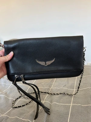 Zadig voltaire  - Stilren svart axelväska i skinn med silverfärgad kedjerem och dekorativt emblem med vingar framtill. Väskan har dragkedja med långa läderdetaljer och en avlång, smidig form som passar perfekt för det viktigaste.