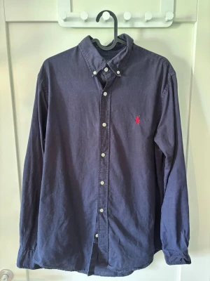 Blå linneskjorta från Ralph Lauren - Klassisk blå skjorta från Ralph Lauren med button-down krage och den ikoniska röda logotypen broderad på bröstet. Skjortan har långa ärmar, vita knappar och är tillverkad i mjuk bomull.