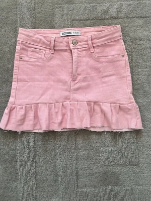 Kort rosa volangkjol från Zara - Supersöt rosa kort volangkjol från Zara med volangkant nertill. Kjolen har klassisk jeansmodell med fickor fram och bak samt bälteshällor. Perfekt för dig som gillar en feminin och trendig stil.