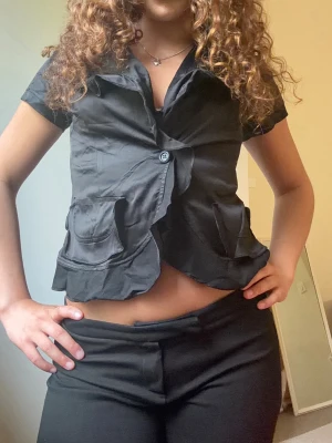 Svart kortärmad blus med fickor - Snygg svart blus med korta ärmar och två stora fickor framtill. Blusen har en knappstängning och vågig kant nertill för en cool look. Tillverkad i ett glansigt tyg som ger en dressad känsla.