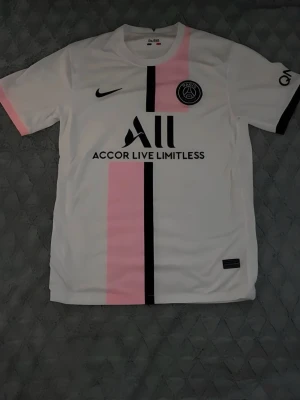 PSG matchtröja vit/rosa Nike - Snygg PSG matchtröja i vitt med rosa och svart rand framtill. Tröjan har rund hals, klubbmärke på bröstet och Nike-logga. Materialet är lätt och andas, perfekt för fotboll eller träning. Stort tryck med 'Accor Live Limitless' på magen.