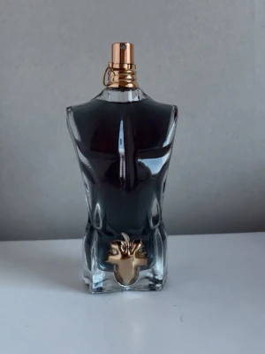125ml Jean Paul Gaultier Le Beau parfym - Jean Paul Gaultier Le Beau parfym i en unik flaska formad som en manlig överkropp. Flaskan är mörkgrön med detaljer i guld, inklusive ett guldfärgat löv framtill. Parfymen levereras i en grön metallburk med gulddetaljer.