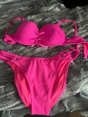 Rosa ribbad bikini med snörning - Snygg bikini i starkt rosa färg med ribbad struktur. Överdelen har vadderade kupor och cool snörningsdetalj framtill. Underdelen har matchande ribbad yta och trendiga band i sidorna. Perfekt för sommarens strandhäng!