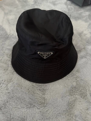 Svart bucket hat från Prada - Stilren svart bucket hat från Prada i glansigt nylonmaterial. Klassisk form med brett brätte och ikonisk metallogo framtill. Perfekt accessoar för en trendig och avslappnad look.