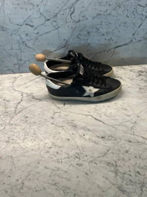 Svarta Golden Goose skor - Snygga svarta sneakers från Golden Goose med vit stjärndetalj på sidan och vit häl. Skorna har svarta skosnören, GGDB-logga på plösen och en klassisk låg siluett. Yttermaterialet är läder och sulan är vit med en något sliten look. Nu pris är 5000!