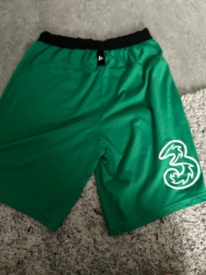Gröna sportshorts från Adidas - Gröna sportiga shorts från Adidas med svart resår i midjan och vit logga på ena benet. Perfekta för träning eller chill. Lätt och bekvämt material som andas.