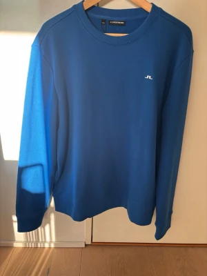 Blå sweatshirt från J.Lindeberg - En stilren blå sweatshirt från J.Lindeberg med rund halsringning och diskret vitt logotryck på bröstet. Tröjan har långa ärmar och ribbade muddar vid ärmslut och nederkant. Perfekt för en avslappnad och snygg look.