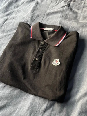 Svart Moncler pikétröja med logga - Svart pikétröja från Moncler med klassisk krage och tre knappar framtill. Kragen har snygga ränder i rött, vitt och blått. Moncler-logga på bröstet ger en exklusiv touch. Perfekt för en clean och stilren look.