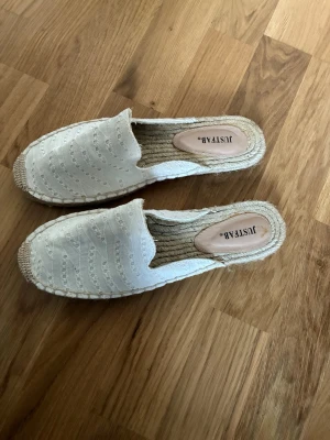 Vita tofflor från JustFab med spets - Säljer ett par vita tofflor från JustFab med snygg spetsdetalj på ovandelen och flätad sula i jute. Perfekta för en avslappnad och trendig look. Lätta att bara slänga på sig och passar till många outfits.