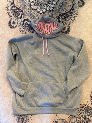 Syna world hoodie - Snygg grå hoodie med rosa dragsko och graffiti-inspirerat tryck i rosa på huvan. Klassisk känguruficka framtill och mjukt material som känns skönt mot huden. Perfekt för dig som gillar streetwear och vill ha en unik detalj i din outfit.