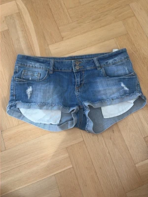 Blå jeansshorts med fransig kant - Snygga blå jeansshorts med fransig kant och slitna detaljer framtill och baktill. Klassisk femficksmodell med dubbla knappar i midjan. Perfekta för varma dagar och en avslappnad stil.