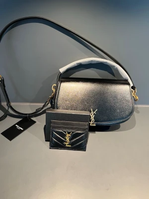 Svart axelväska från Saint Laurent - Saint Laurent väska i nyskick. Top kvalitet med fina detaljer och väldigt exklusiv känsla. Väskan kommer med dustbag, tags och cardholder som på bilderna. Kan användas både som shoulder bag och crossbody tack vare den justerbara remmen.  Väldigt clean och elegant modell som passar till allt. Inga skador eller slitningar, helt ny.