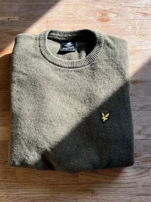 Olivgrön stickad tröja Lyle & Scott - Snygg olivgrön stickad tröja från Lyle & Scott med rund halsringning och klassisk gul logga broderad på bröstet. Tröjan har ribbade muddar vid hals, ärmslut och nederkant. Perfekt för lager-på-lager och stilren vardagslook.
