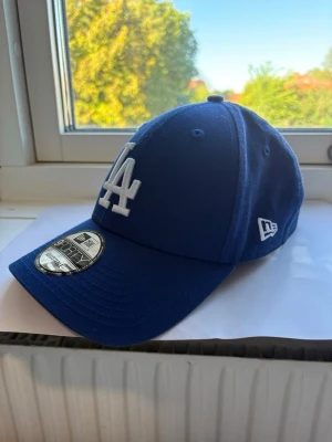 Blå LA Dodgers keps från New Era - Säljer en blå keps från New Era med LA Dodgers-logga broderad i vitt framtill. Klassisk böjd skärm och justerbart spänne bak. Kepsen är tillverkad i bomull och har ventilerande hål upptill för extra komfort.