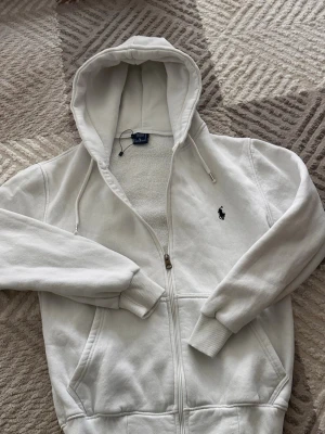 Vit hoodie från Polo Ralph Lauren - Klassisk vit hoodie från Polo Ralph Lauren med dragkedja framtill och broderad svart logga på bröstet. Hoodien har huva med dragsko, ribbade muddar och fickor på sidorna. Tillverkad i mjuk bomullsblandning för extra komfort.