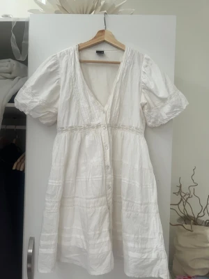 Vit kort klänning från Gina Tricot - Superfin vit klänning från Gina Tricot med v-ringning, korta puffärmar och knappar framtill. Klänningen har fina spetsdetaljer och är tillverkad i ett lätt bomullstyg som passar perfekt till varmare dagar.