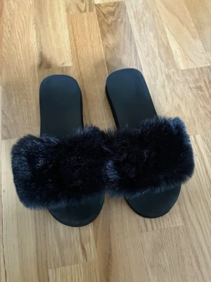 Svarta fluffiga tofflor med fuskpäls - Säljer ett par svarta tofflor med bred rem i mjuk och fluffig fuskpäls. Sulan är platt och i svart material, perfekt för en chill och trendig look hemma eller på stan. Märke ej angivet.