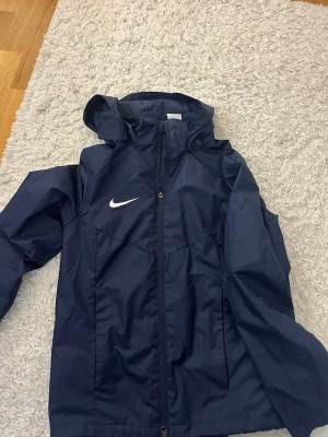 Mörkblå Nike vindjacka med huva - Snygg mörkblå vindjacka från Nike med vit logga på bröstet. Jackan har dragkedja framtill, huva och två sidofickor. Perfekt för blåsiga dagar och sportiga outfits. Materialet är lätt och vattenavvisande.
