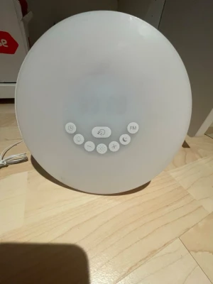 Vit wake up light med FM-radio - Rund vit väckarklocka med digital display, flera knappar för funktioner som FM-radio, ljus och alarm. Kan drivas med sladd eller batterier. Perfekt för att vakna till ljus och ljud. Modern design i plast, enkel att använda.