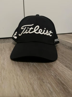 Svart Titleist keps med brodyr - Snygg svart keps från Titleist med vit broderad logga framtill och texten 'PRO' på sidan. Kepsen har böjd skärm och mesh-paneler bak för extra ventilation. Perfekt accessoar för en sportig och stilren look. Pris kan absolut diskuteras.   