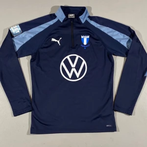 Helt ny Malmö FF träningströja Puma blå (FLERA STORLEKAR)  - Mörkblå långärmad träningströja från Puma med Malmö FF-logga på bröstet och Volkswagen-tryck på magen. Tröjan har ljusblå detaljer på axlarna, halv dragkedja och är tillverkad i funktionsmaterial. Perfekt för fotbollsträning eller sportiga aktiviteter.
