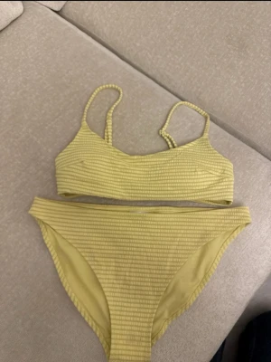 Gul ribbad bikini med smala axelband - Trendig gul bikini med ribbad struktur och smala axelband. Överdelen har en enkel, sportig form och bikinitrosan är högt skuren. Perfekt för strandhäng eller poolparty. Materialet känns mjukt och följsamt mot huden.